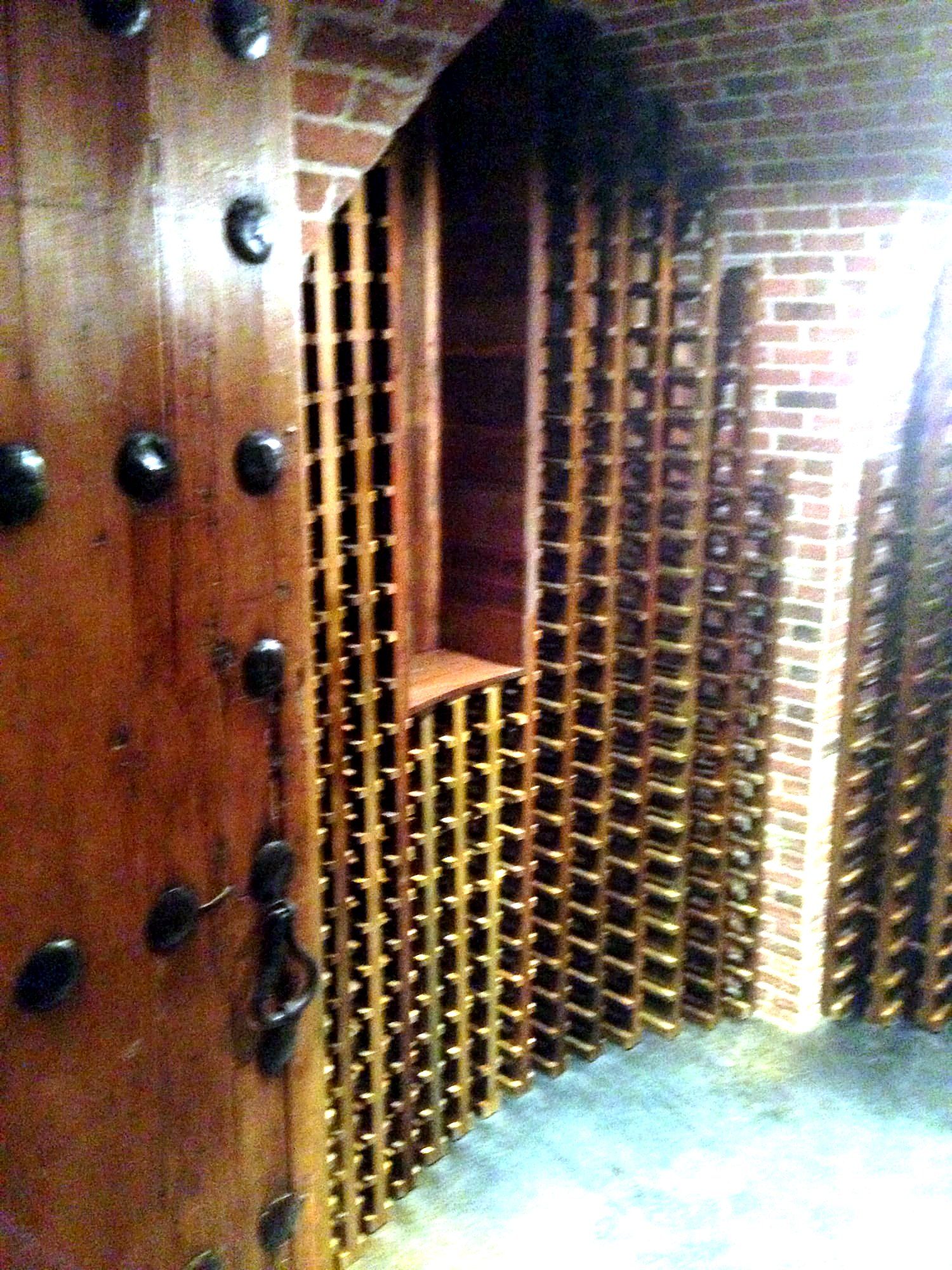 Dark wood cellar door with wrought iron strap hinges opening onto a brick-lined wine cellar with rows of diagonal wood wine racks