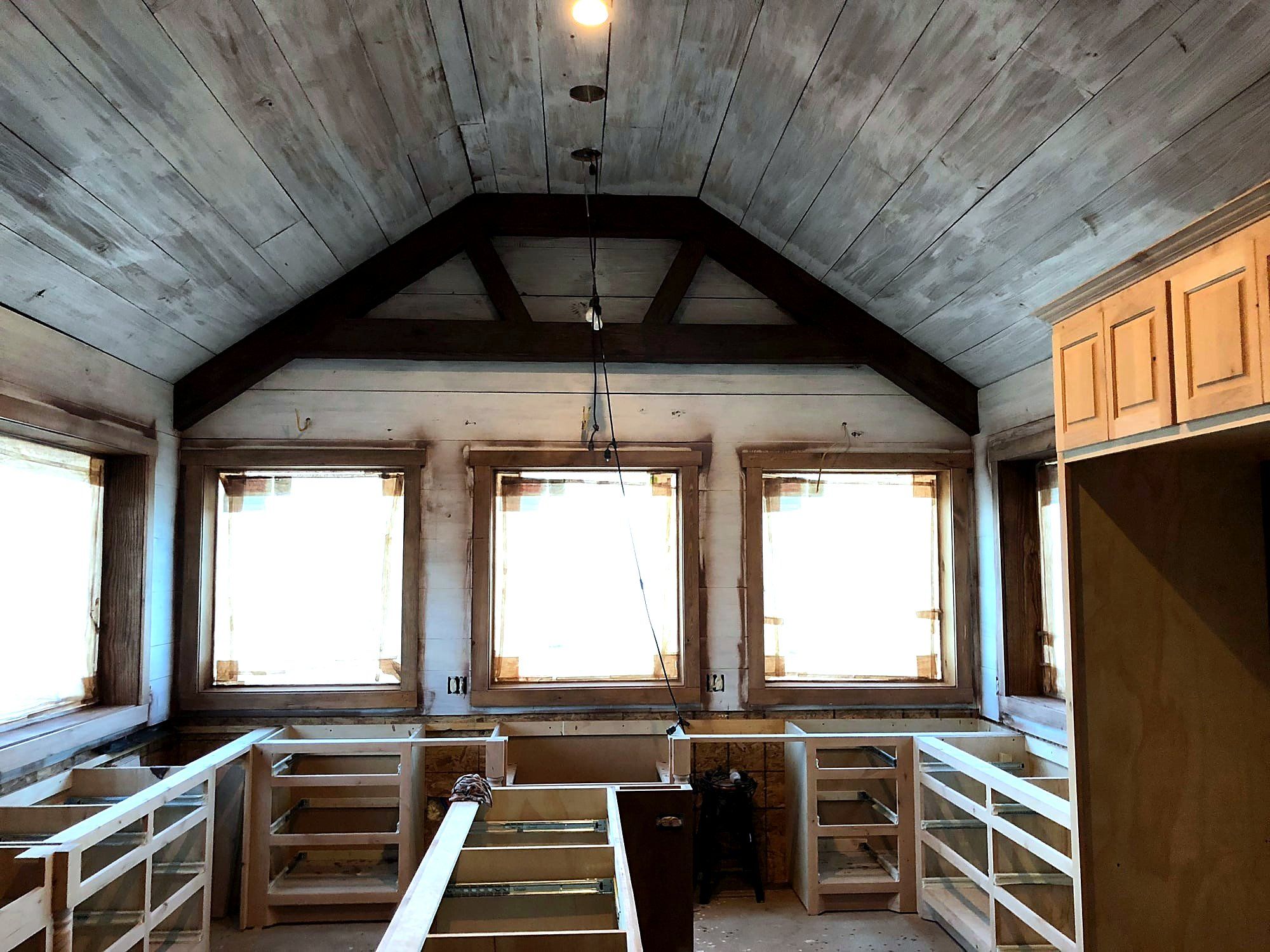 Vaulted great room ceiling with whitewashed tongue-and-groove boards framed by a dark stained king-post truss, cabinetry not yet countertop-ready below