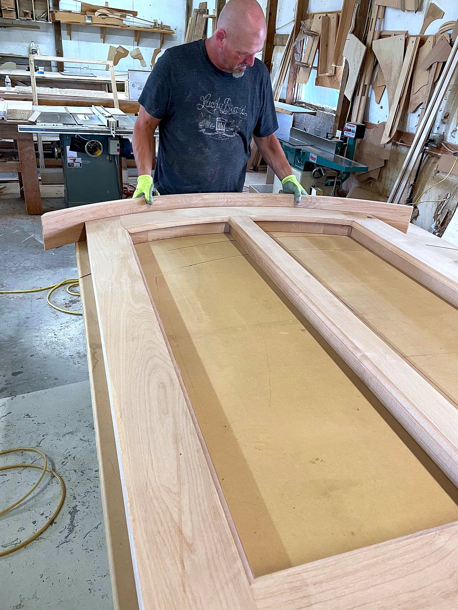 Craftsman supporting a curved-top door frame with divided lites on a shop bench, checking the fit of the arched rail