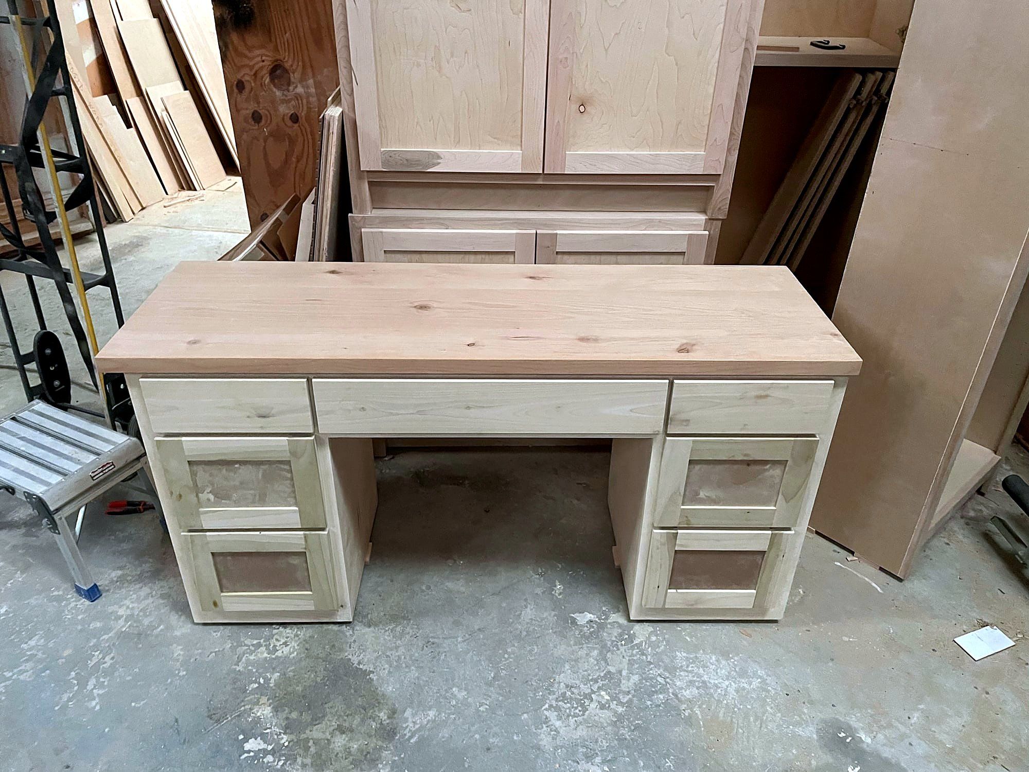 Kneehole pedestal desk with paneled drawer fronts and a banded top, natural finish, staged in the shop in front of a large armoire